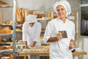 Girl works as baker in bakery, makes bakery products from dough. Worker created lump of dough from ingredients, prepares it for cutting. She is standing in work area of bakery shop production