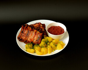 Baked pork ribs and boiled potatoes with dill and tomato sauce on a black background.