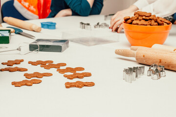 A baking scene with gingerbread cookies shaped like people, metal cutters, a rolling pin, and an orange bowl filled with cookies. tools and hands suggest active participation in a festive kitchen act.