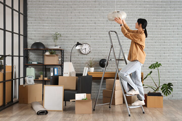 Young businesswoman with ladder and cardboard boxes packing lamp in office on moving day
