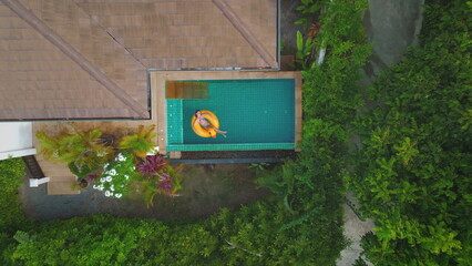 Aerial view of a woman enjoying a relaxing day by floating on a yellow inflatable ring in a rooftop pool, surrounded by the lush greenery of a tropical forest