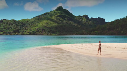 Young woman is enjoying a sunny day, walking on a white sand beach with turquoise water and lush green mountains in the background