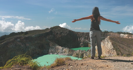 Obraz premium Woman raise hands on volcano mountain crater edge. Volcanic Kelimutu blue crater lakes. Travel destination. Beautiful wild landscape. Female tourist on summer vacation. Lifestyle holiday concept