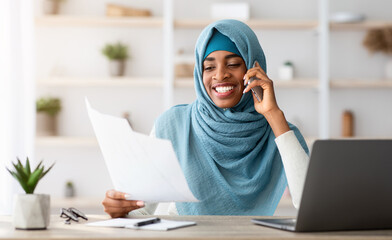 Young Black Muslim Businesswoman Talking On Cellphone And Working With Papers In Office, Islamic Female Entrepreneur Sitting At Desk With Laptop, Checking Financial Documents And Speaking On Phone