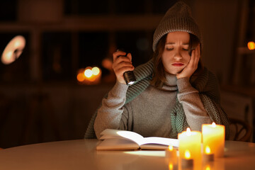 Young woman with flashlight reading book at table in kitchen during blackout