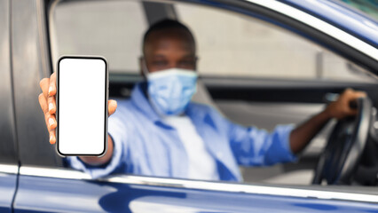 Closeup of black man in surgical mask showing mobile phone with white empty screen for mock up sitting on driver seat in car, holding gadget in hand, selective focus on device, covid protection