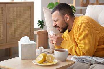 Ill young man sneezing near lemon tea on sofa at home