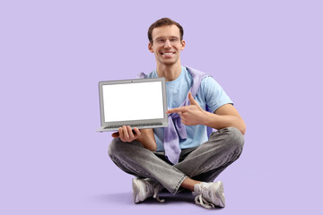 Young man pointing at blank laptop on lilac background