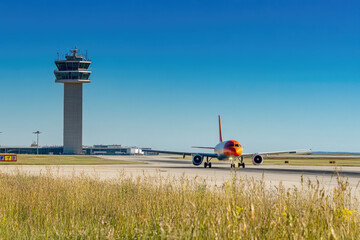 Airplane taxiing on the runway, with airport control tower in view. Copyspace on the left. Bright, sunny lighting. Clear blue sky and runway background.