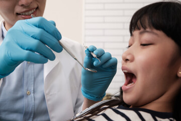 A young Asian male pediatric dentist checks and examines a little Thai girl's teeth in dental clinic, well-being mouth oral hygiene, and professional orthodontic healthcare work in a kid hospital.