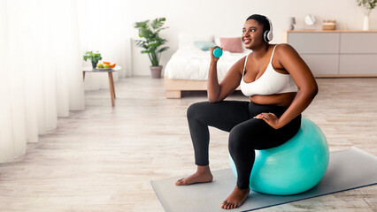 Obraz premium Overweight Afro woman exercising with dumbbells, sitting on fitball, listening to music in headphones indoors, copy space. Curvy black lady doing home fitness, losing weight, full length