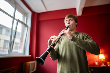 Clarinet player. Young man clarinetist playing flute woodwind music instrument at home with red wall, soft focus. Musical instruments. Guy plays the oboe in education classroom. Hobby. 