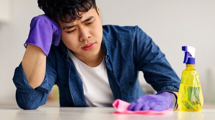 Exhausted young asian man leaning on his hand, wearing gloves, holding dust cloth, having break...