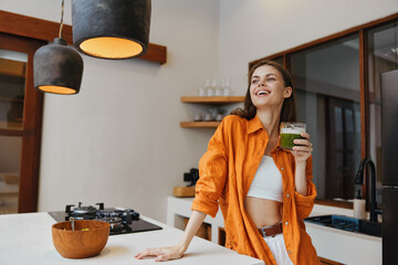 Young woman enjoying a healthy green smoothie in a bright kitchen, wearing an orange shirt and smiling, reflecting wellness and vitality