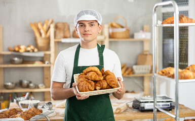 Positive male baker in uniform stands behind counter in small private bakery offering hot croissants
