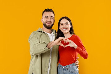 Obraz premium A young couple stands together against a vibrant yellow backdrop, smiling and making a heart shape with their hands. Their affectionate pose reflects their love and happiness.