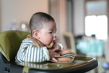 A six-month-old Chinese boy sits on a chair and eats a bib on a cold winter day in an apartment...