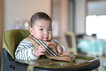 A six-month-old Chinese boy sits on a chair and eats a bib on a cold winter day in an apartment...
