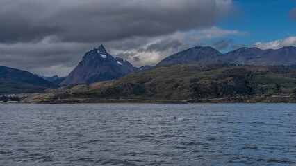 Mountains with snow-capped peaks against a blue sky, clouds. The road along the ocean shore. The city of Ushuaia by the sea. The tail of a whale is visible above the surface of the water. Beagle Canal