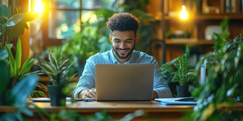 Happy businessman writing in diary while working on laptop in modern office surrounded by green plants