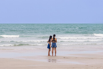 Back view of happy carefree women using mobile phone at beach. Woman friends using smartphone at seaside. Beautiful lovely two women spending time at beach. Concept of travel, tourism and vacation.