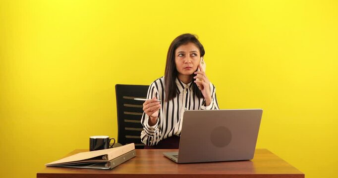 Young woman sitting at desk with laptop talking on smart phone isolated over yellow background.