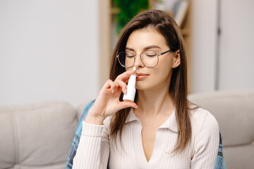 Young woman wearing casual clothing, using a nasal spray to facilitate breathing, sitting on a sofa...