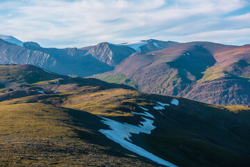 Scenic sunny aerial top view to sunlit big green hills against bright high rocky mountain range in bright sun under clouds in blue sky. Light and shadows on colorful large mountains just before sunset