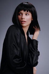 A woman with dark hair, dressed in a black jacket, stands against a plain gray backdrop, looking directly at the camera with a confident expression
