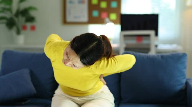 Woman is sitting on a sofa and experiencing back pain. She is massaging her lower back with a pained expression.