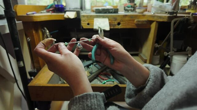 A female jeweller holds a bracelet and a pair of snips at her workbench during the day.
