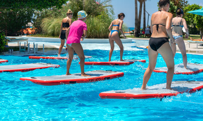 A group of women performing yoga poses on paddleboards in a bright resort pool, surrounded by tropical greenery. Concept of unique fitness activities and wellness-focused vacations