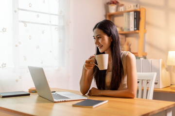 Drinking coffee, asian woman working on laptop at home office desk, copy space
