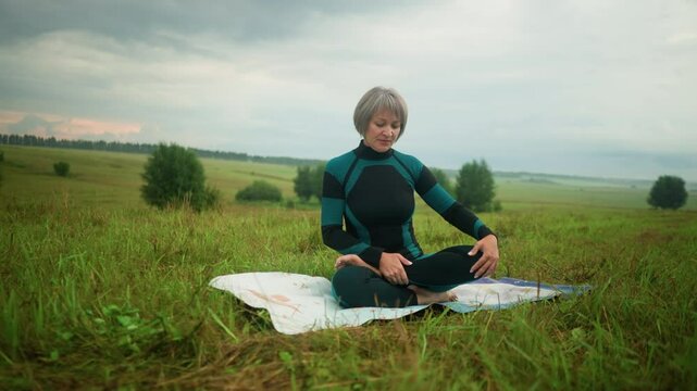 Woman seated on mat in siddhasana pose practicing yoga with calm focus, yellow boot and green raincoat visible in the background, set against a vast grassy field under a cloudy sky