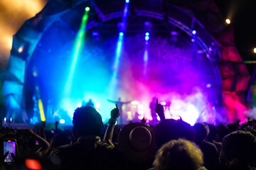 Silhouettes of crowd at concert in front of stage with bright spotlights.The back view of people enjoy live music at the band's performance. Outdoor concert with musicians on stage and fans.