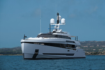 A huge, innovative white yacht is anchored off the coast of Sardinia. The mega yacht rests on blue water, with the mountainous relief and blue sky in the background.