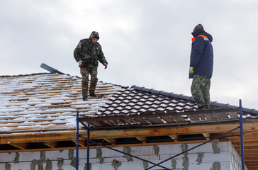 Two men are working on a roof, one of them wearing a blue jacket