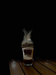 Glass Cup of Coffee on Wooden Table in Dark Minimalist Setting. A simple composition of a glass coffee cup with a blurred carafe in the background, illuminated softly in a dark environment