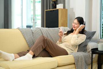 Fototapeta premium Relaxed young asian lady in wireless headphones using smartphone on sofa