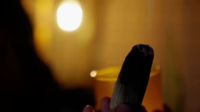 Shaman holds a smoking palo santo stick, surrounded by a dark, blurred background. Warm light creates a calming atmosphere, enhancing the spiritual and healing essence of the ritual