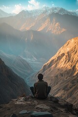 Fototapeta premium A lone figure sits in contemplation, overlooking a vast mountain landscape under a clear, blue sky at sunset.