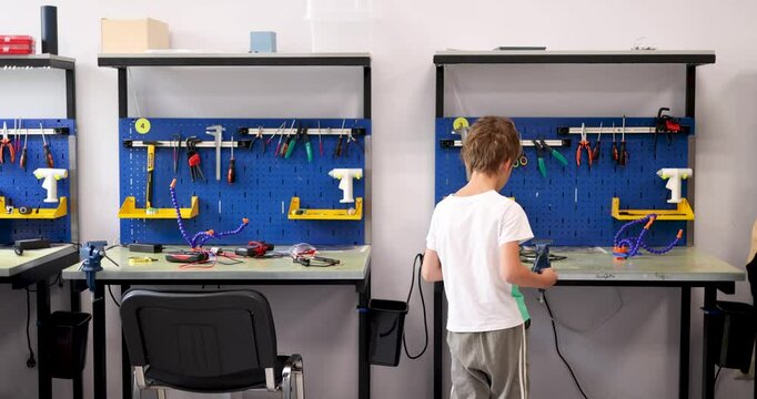 Real time handheld shot back view of faceless boy in casual clothes looking down while standing near counter in workshop along turning moveable bench vice with attached rod