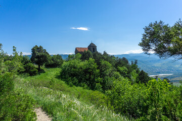 Fototapeta premium The small church on the hill among bushes and trees is part of the Shio-Mgvime monastery. The bright blue sky, river and mountains in the background.