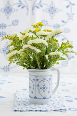 White and yellow flowers in a patterned ceramic pitcher on a blue background