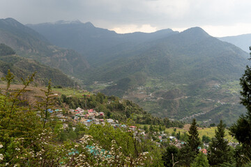 A breathtaking view of a mountain valley with scattered settlements, lush greenery and terraced fields. Snow-capped peaks rise in the distance under an overcast sky, creating a serene landscape.