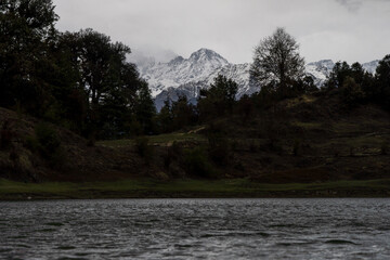 A serene lake with rippling water at the foreground, surrounded by rugged terrain and dense trees. Snow-capped mountains rise dramatically in the background under a dark and cloudy sky at deoria lake.