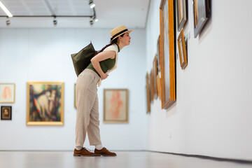 Wide shot of a young Caucasian woman in a straw hat and elegant clothes carefully contemplating modern art in the gallery. Low angle side view. The concept of the Museum Day