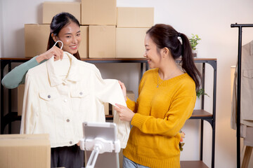 Two young asian woman live online clothing store while showing clothing with tablet on social at home, entrepreneur streaming for selling online clothing and presenting, online seller.