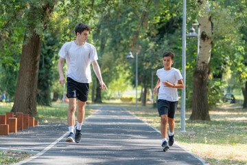Trainer runs alongside two boys in a sunny park, focusing on improving their endurance and physical fitness. The boys are energized, learning proper running techniques while enjoying outdoor exercise