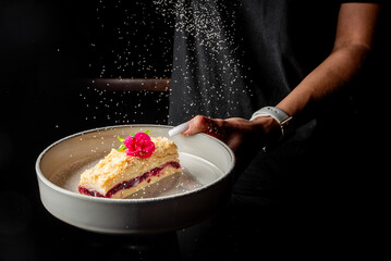 A slice of vibrant layered dessert is elegantly presented on a white plate, garnished with fresh flowers, as fine sugar is sprinkled overhead against a dark backdrop.
